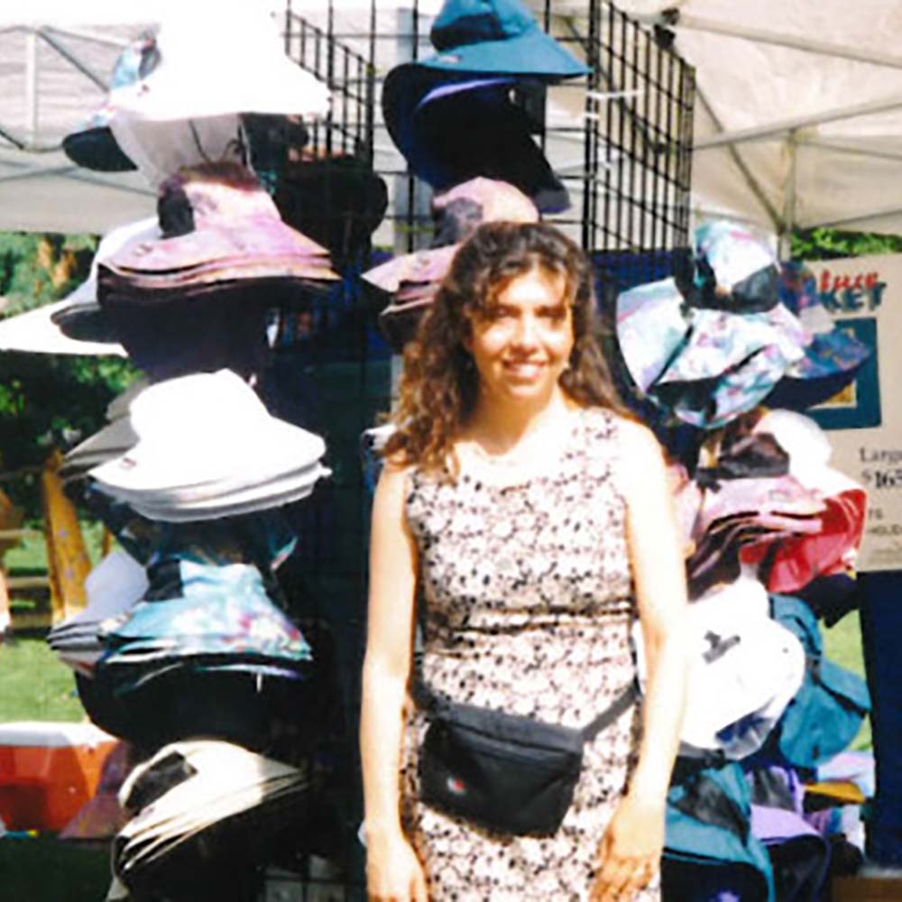 A photo of Angeline Lacy in front of a rack of Sunday Afternoons hats at a craft show booth.