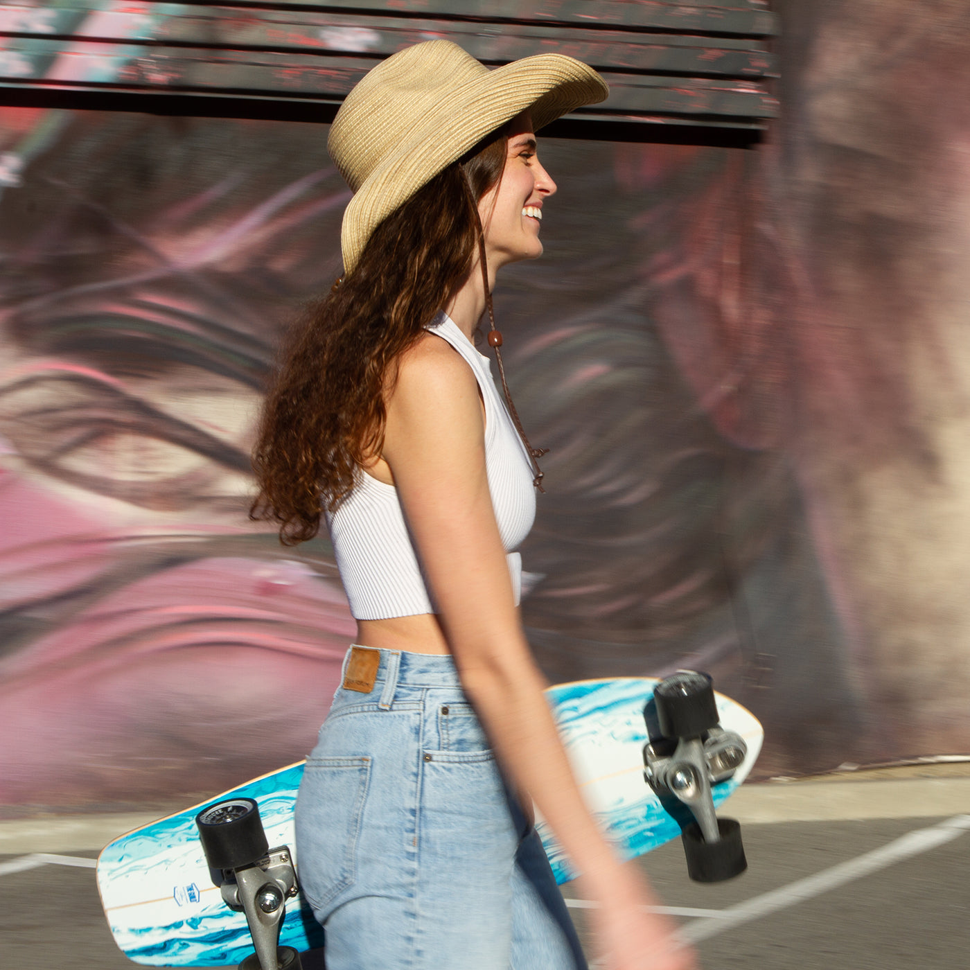 A woman walking, holding a longboard, and wearing a Sunday Afternoons Sunset Hat, in an urban setting.